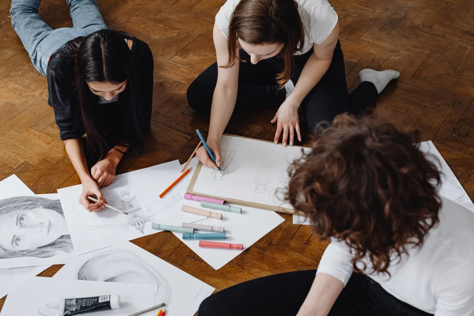 Three women engage in a collaborative drawing session on a wooden floor, surrounded by sketches and art supplies.